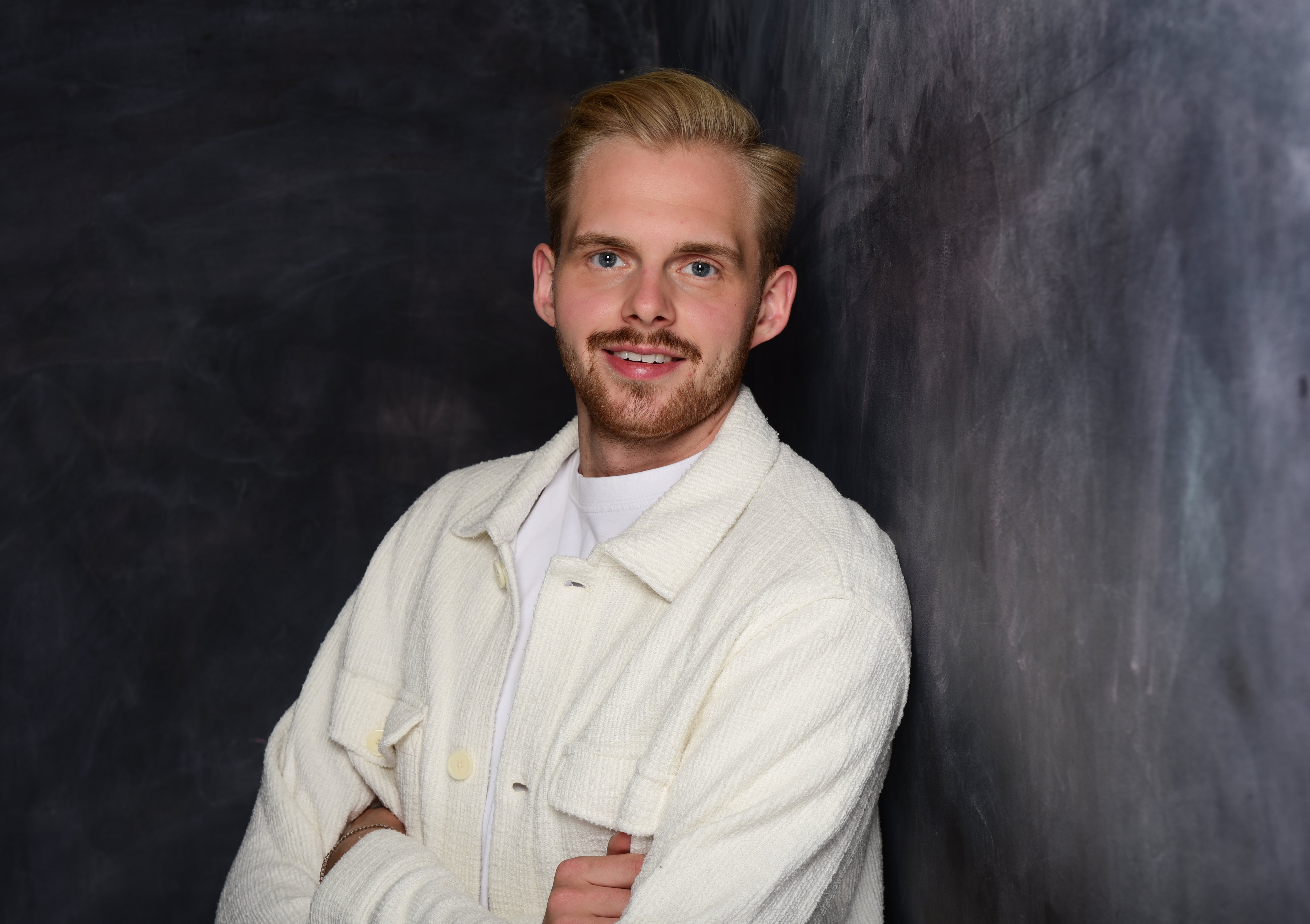 Portrait of Luca Büürma smiling with short blond hair and light beard, wearing a white textured jacket and white t-shirt, standing against a dark chalkboard background.
