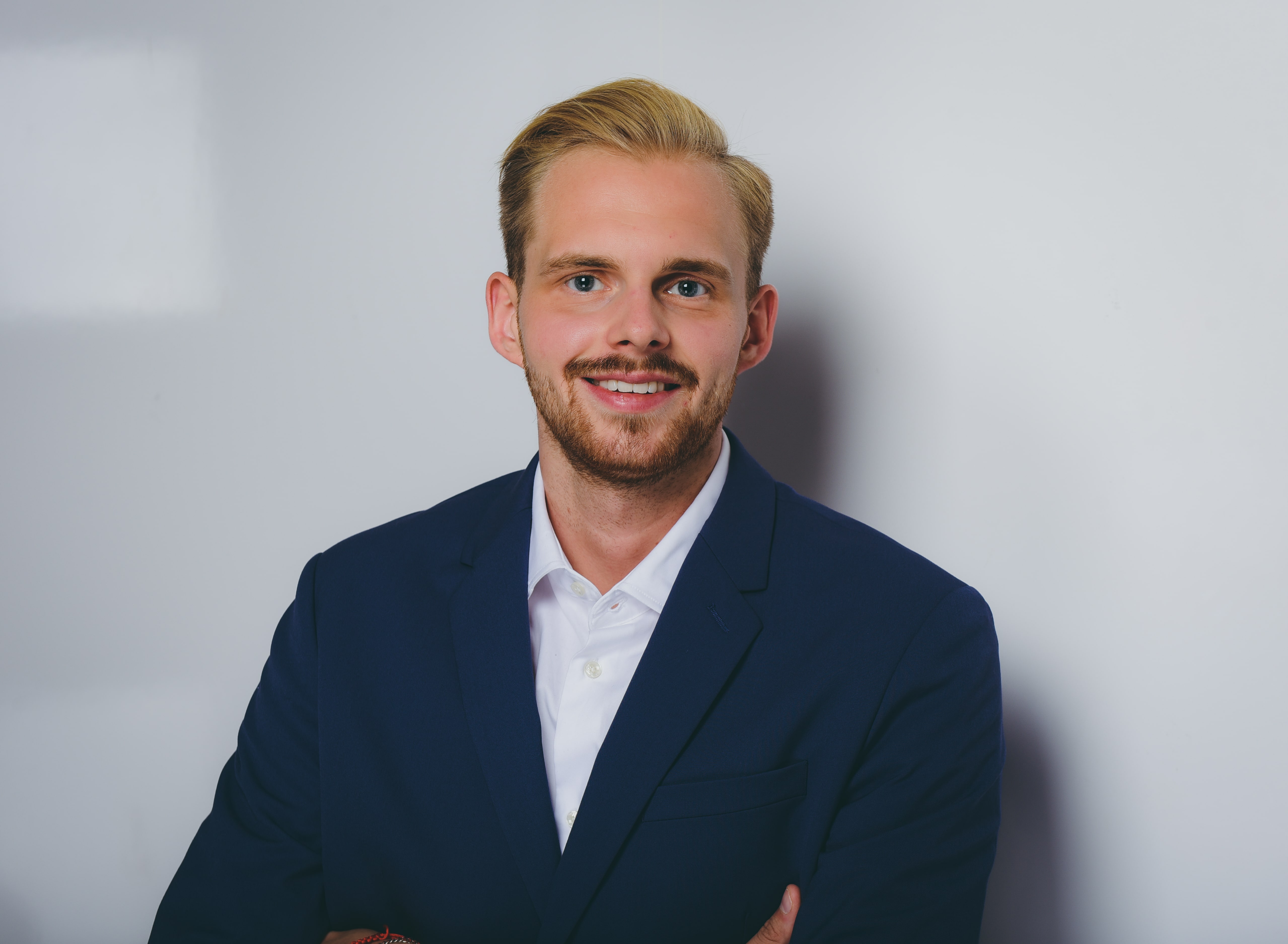 Portrait of Luca wearing a blue suit and white shirt, with short blond hair, smiling in front of a white background.
